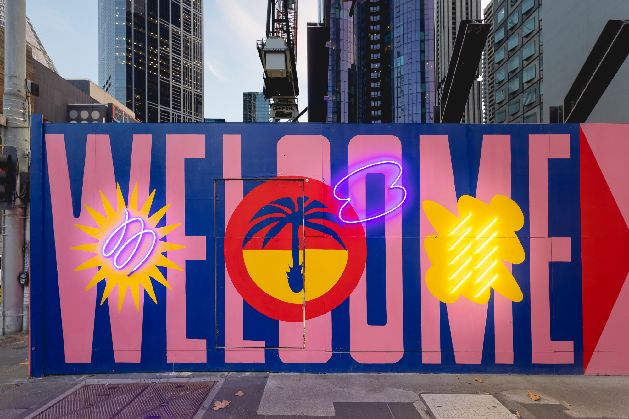 Artwork on a temporary construction fence says 'welcome' with neon lights in front of skyscrapers