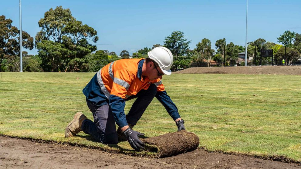 A construction worker wearing protective clothing and hard hat laying natural turf on the Greensborough College oval.