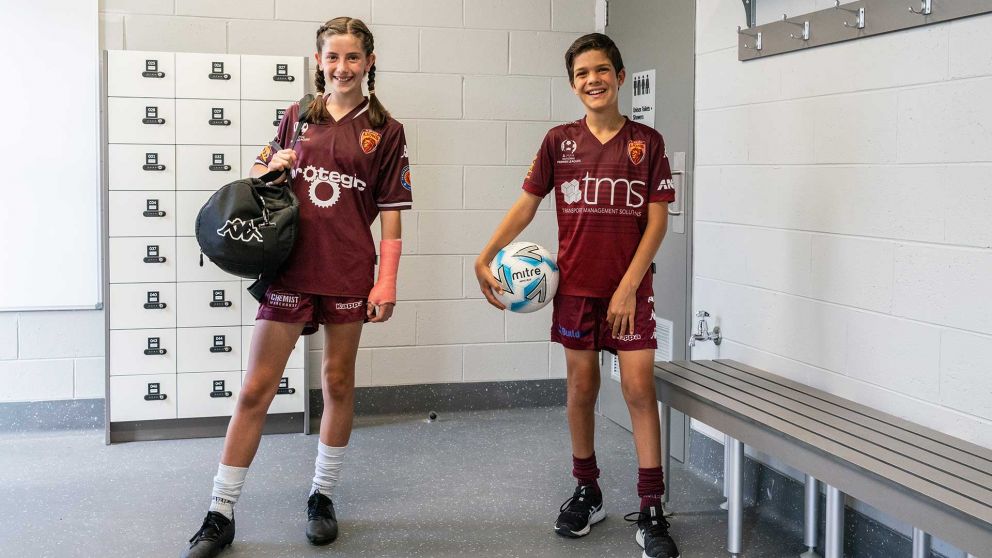 Two young FC Bulleen Lions players in the Veneto Club locker room soccer. One of them is holding a soccer ball and the other is carrying a large sports bag. 