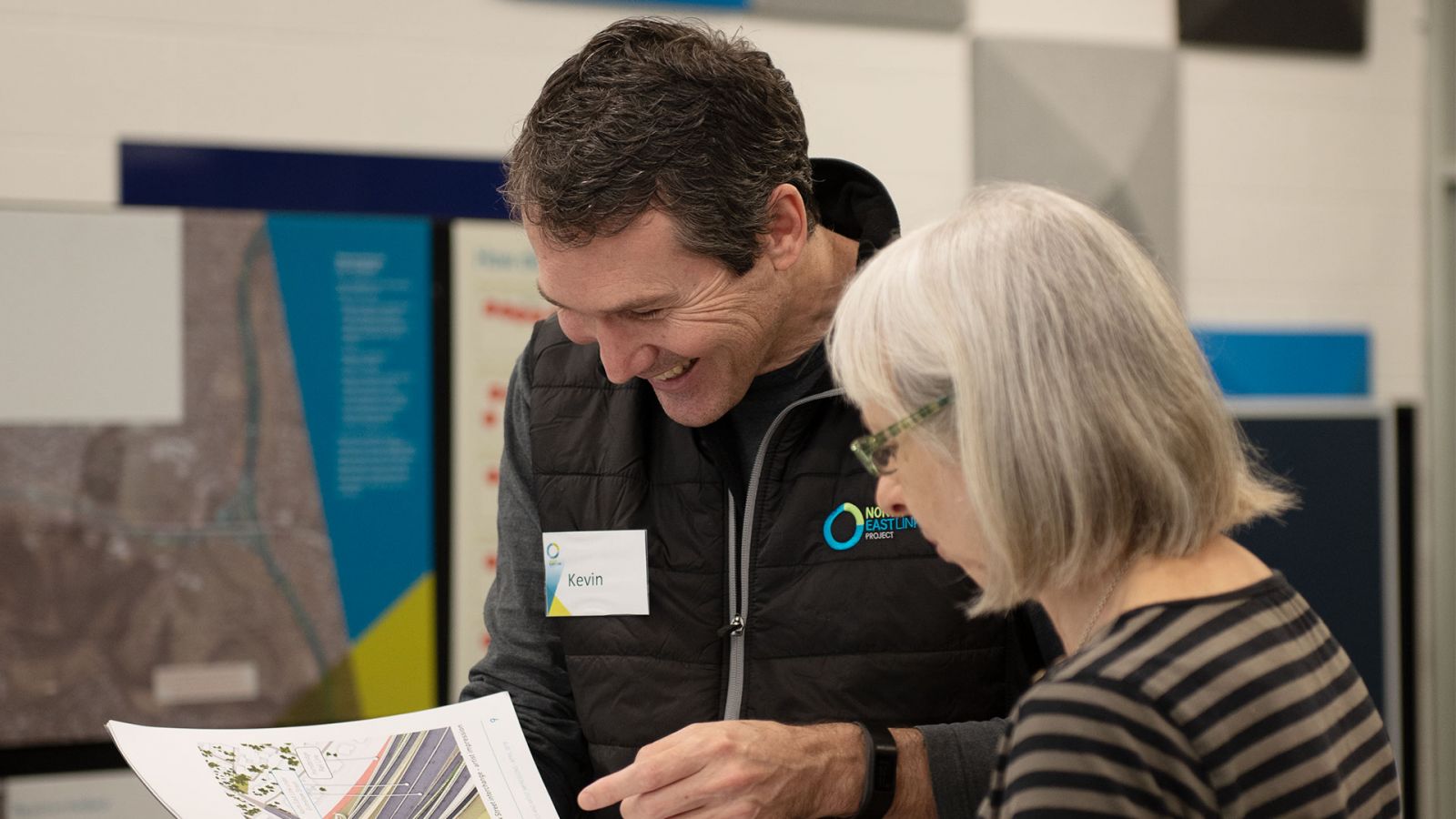 A man and woman standing and looking at a document together with large posters in the background. The man is wearing a black vest with the North East Link Project logo on it and he is smiling, the woman is wearing glasses. 