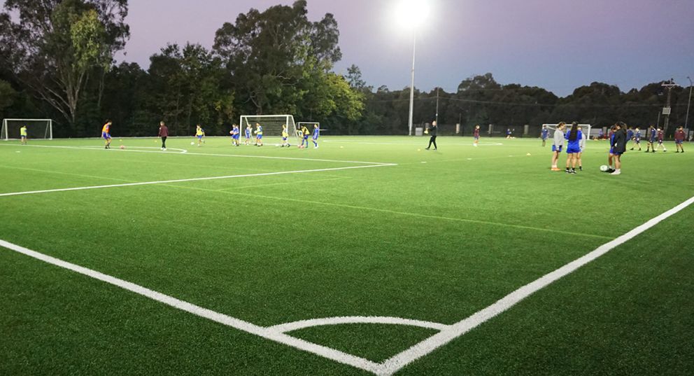 A wide shot of the synthetic soccer pitch at Veneto Club at dusk. The floodlights are turned on and numerous players are practising soccer in groups.