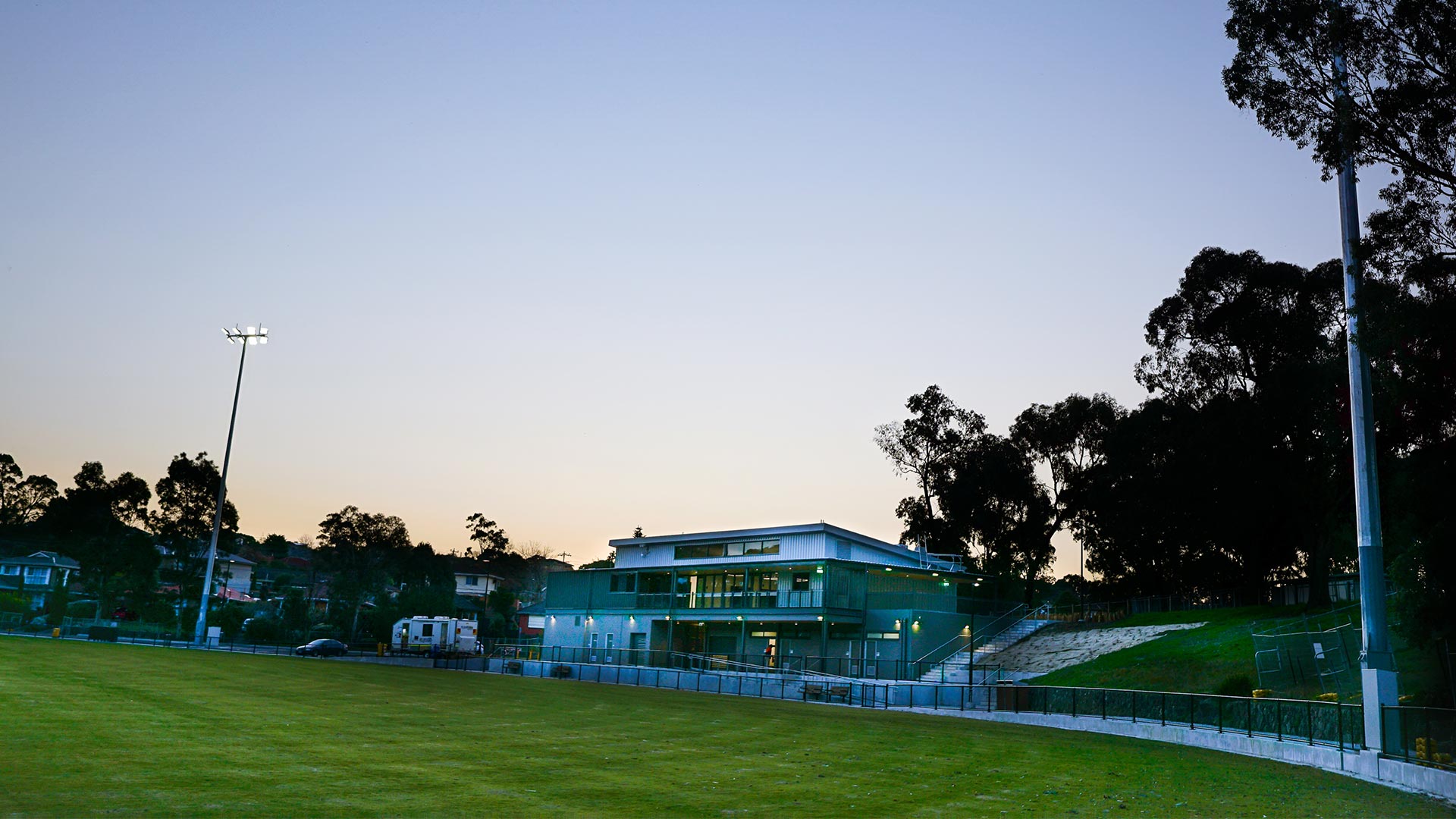 Looking at Binnak Park pavilion and car park from the oval at dusk, surrounded by mature trees to the right and houses to the left. The interior and exterior pavilion lights are turned on and the oval floodlights are also turned on.