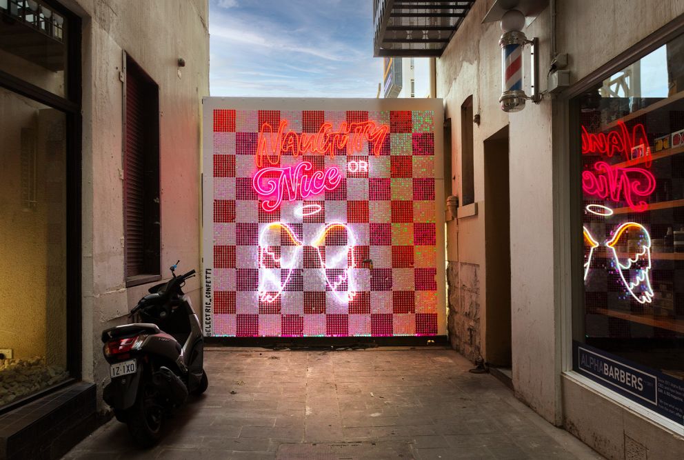 A pink, red and white chequered wall with neon angel wings and writing that says 'Naughty or nice'.