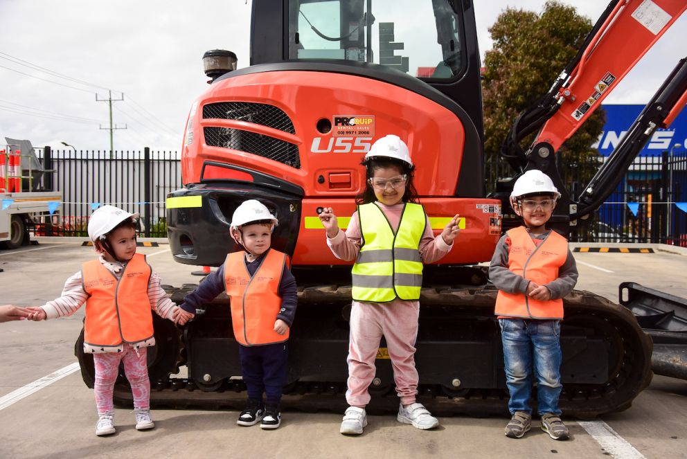 Four young children in hi-vis clothing stand in front of a digger.