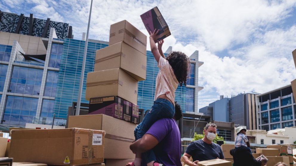 A child is lifted up by an adult reaching out to put a cardboard box on the top of a stack of boxes.