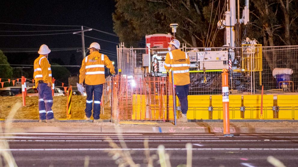 Three construction workers wearing high-vis clothing and protective equipment standing at a work site at night. There is heavy machinery behind temporary fencing and bright yellow safety barriers.