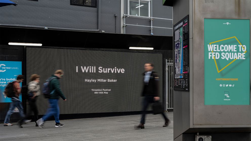 Image of people walking past a digital screen on the side of a construction site. Text on screens reads 'I Will Survive'