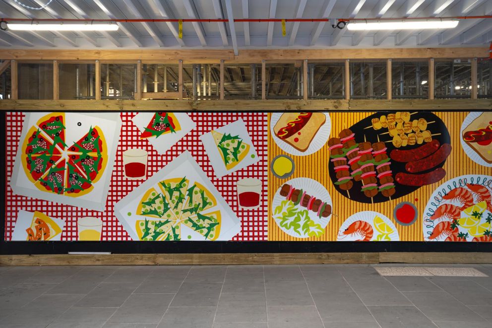 An artwork featuring an assortment of colourful foods on patterned tablecloths installed on a construction hoarding