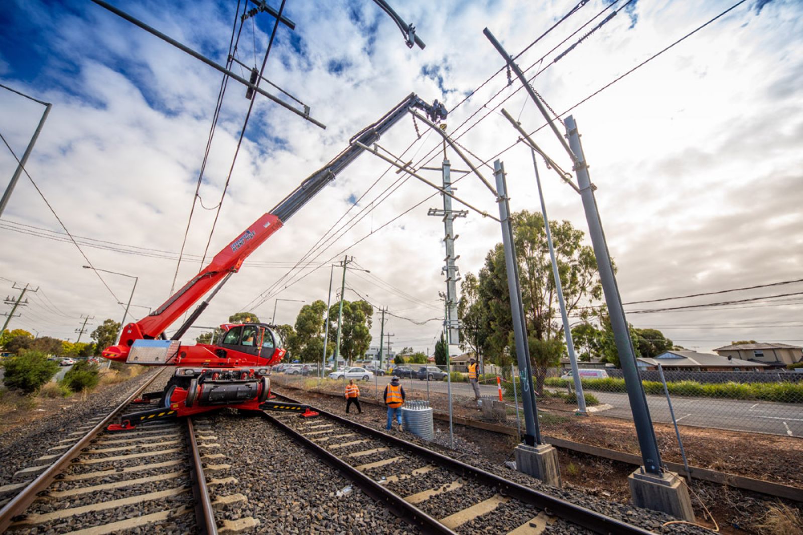A crane sits on the rail track while workers oversee upgrades to the overhead wiring structures.