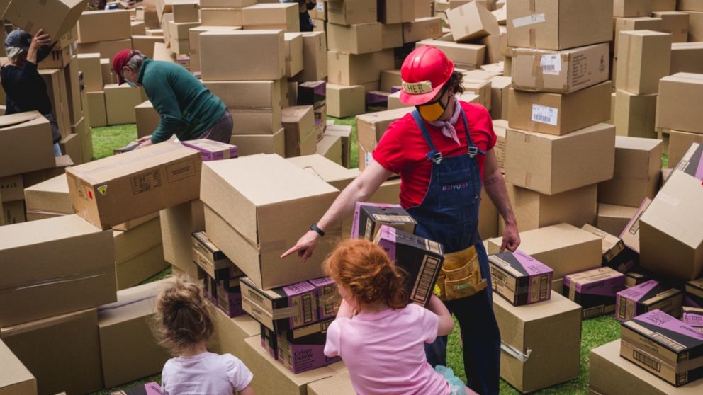 A staff dressed in red helps two small children who are playing with cardboard boxes.