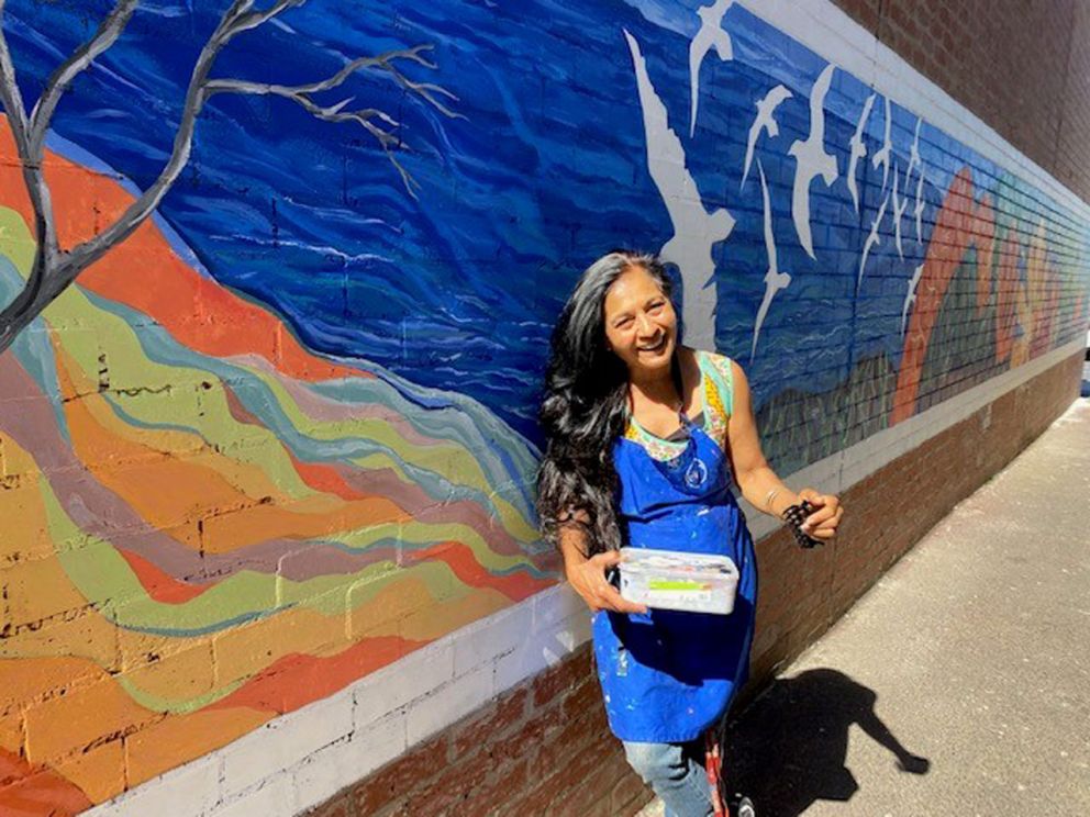 Artist Anu standing in front of a long wall of artwork.