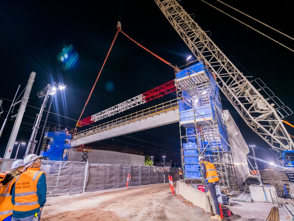Workers watching a large concrete beam being lifted in the air at night.