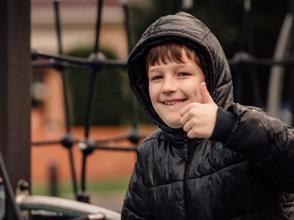 A boy in.a hooded puffer jacket giving a big thumbs up while on Burwood Park playground. 