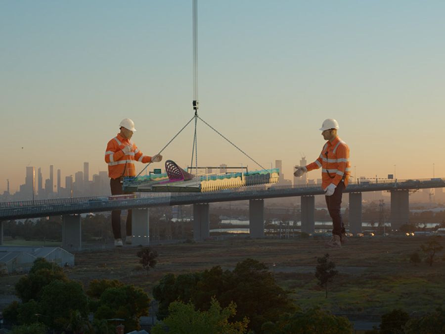 Two giant people in safety clothing stand either side of the construction of a raised road in front of the Melbourne city skyline at sunset.
