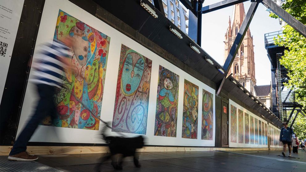 2 figures walking past colourful construction hoarding, one of them holding an umbrella. Artwork includes a row of colourful, abstract portraits.