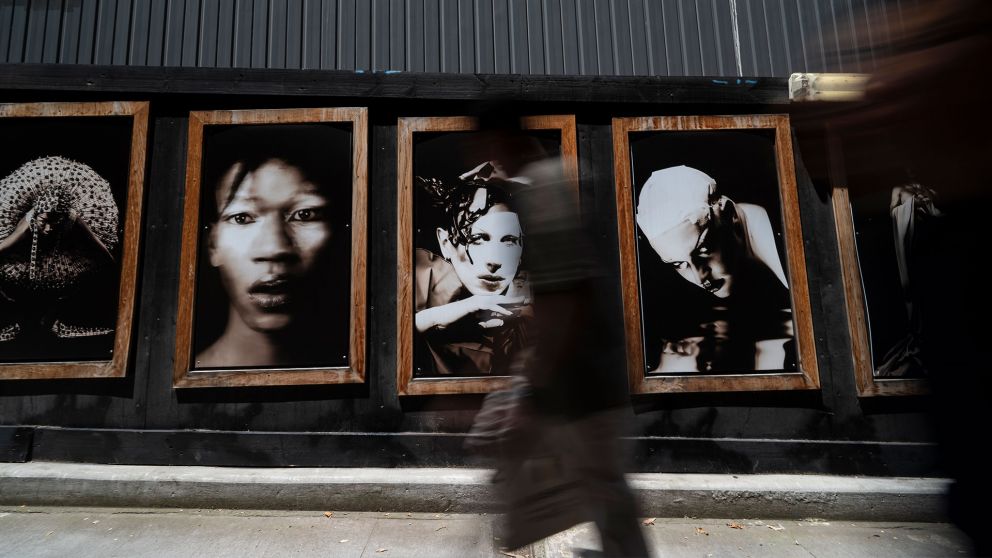 Four wooden frames with black and white portraits and two blurred figures walking past.