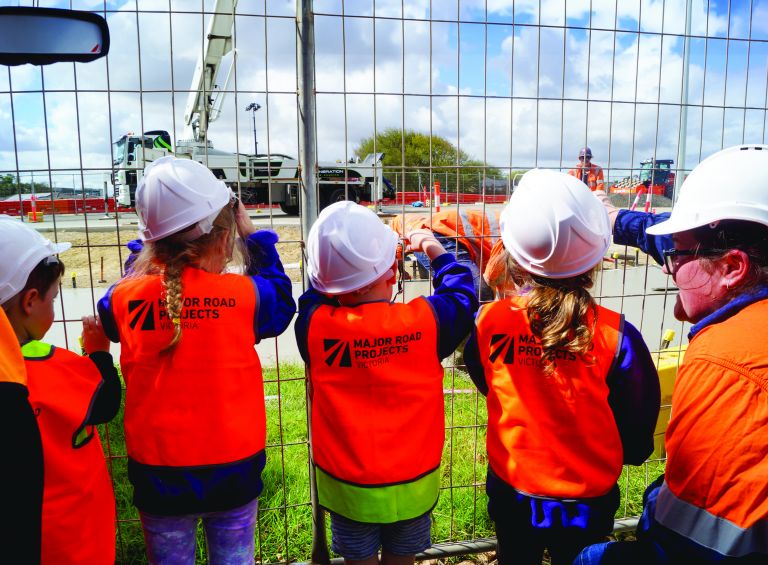 3 children and an adult dressed in high-vis facing with their backs showing looking beyond a fence where construction is happening