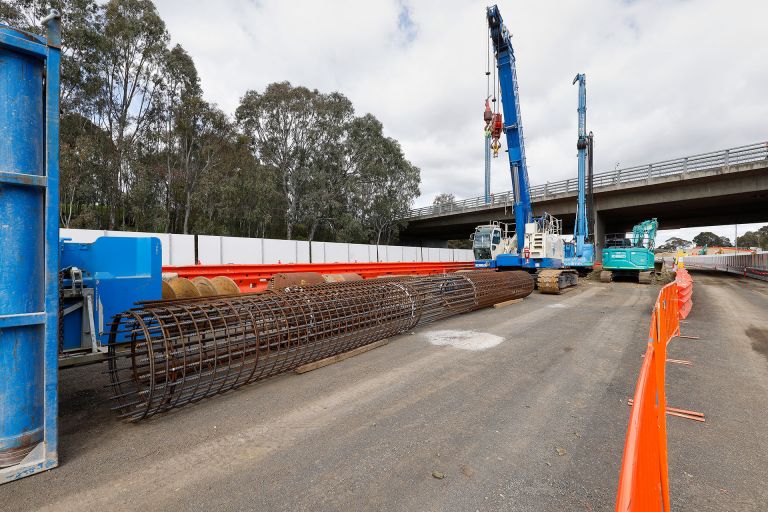 A construction site featuring large steel reinforcement cages for concrete piles laid on the ground. Two blue heavy-duty piling rigs are positioned in the background. The site is bordered by orange safety barriers, and an overpass bridge runs above the scene.