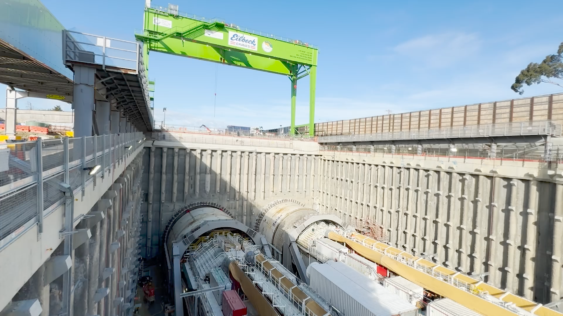 A large construction site featuring two massive tunnel boring machines (TBMs) situated within a deep excavation pit, surrounded by reinforced concrete walls. Above the site, a bright green gantry crane with 'Edilcek Cranes' branding is suspended, indicating active construction work. The scene shows extensive scaffolding and machinery for tunneling, under clear blue skies.