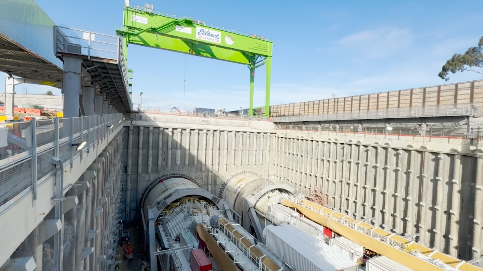 A large construction site featuring two massive tunnel boring machines (TBMs) situated within a deep excavation pit, surrounded by reinforced concrete walls. Above the site, a bright green gantry crane with 