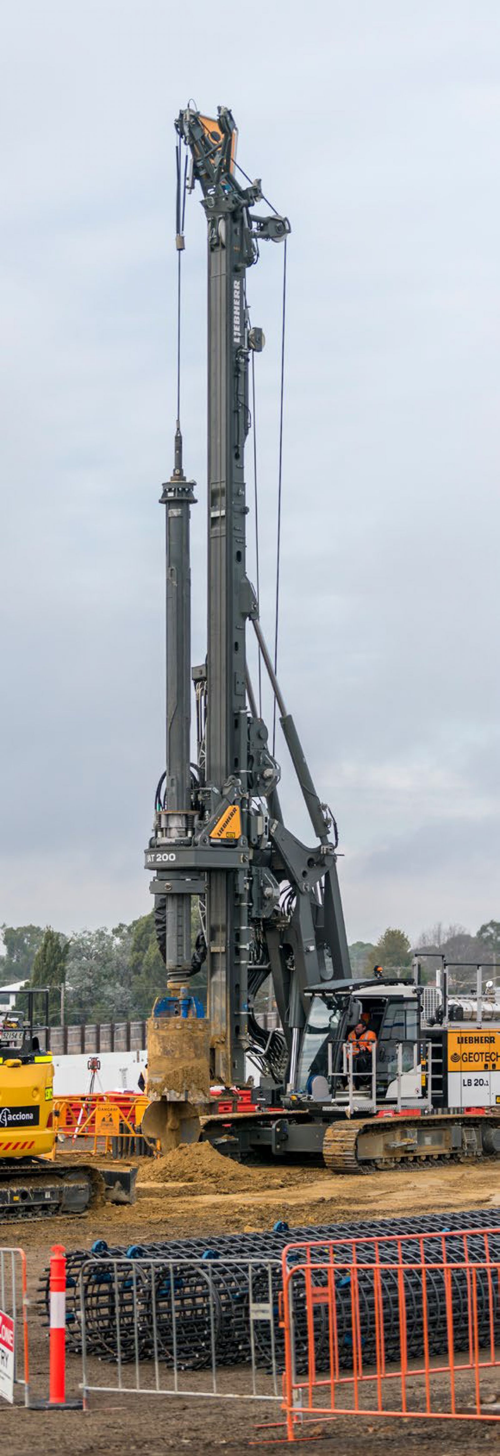 A large drilling rig in operation at a construction site, with a worker controlling the machine as it digs into the ground, surrounded by rebar stacks and safety fencing.