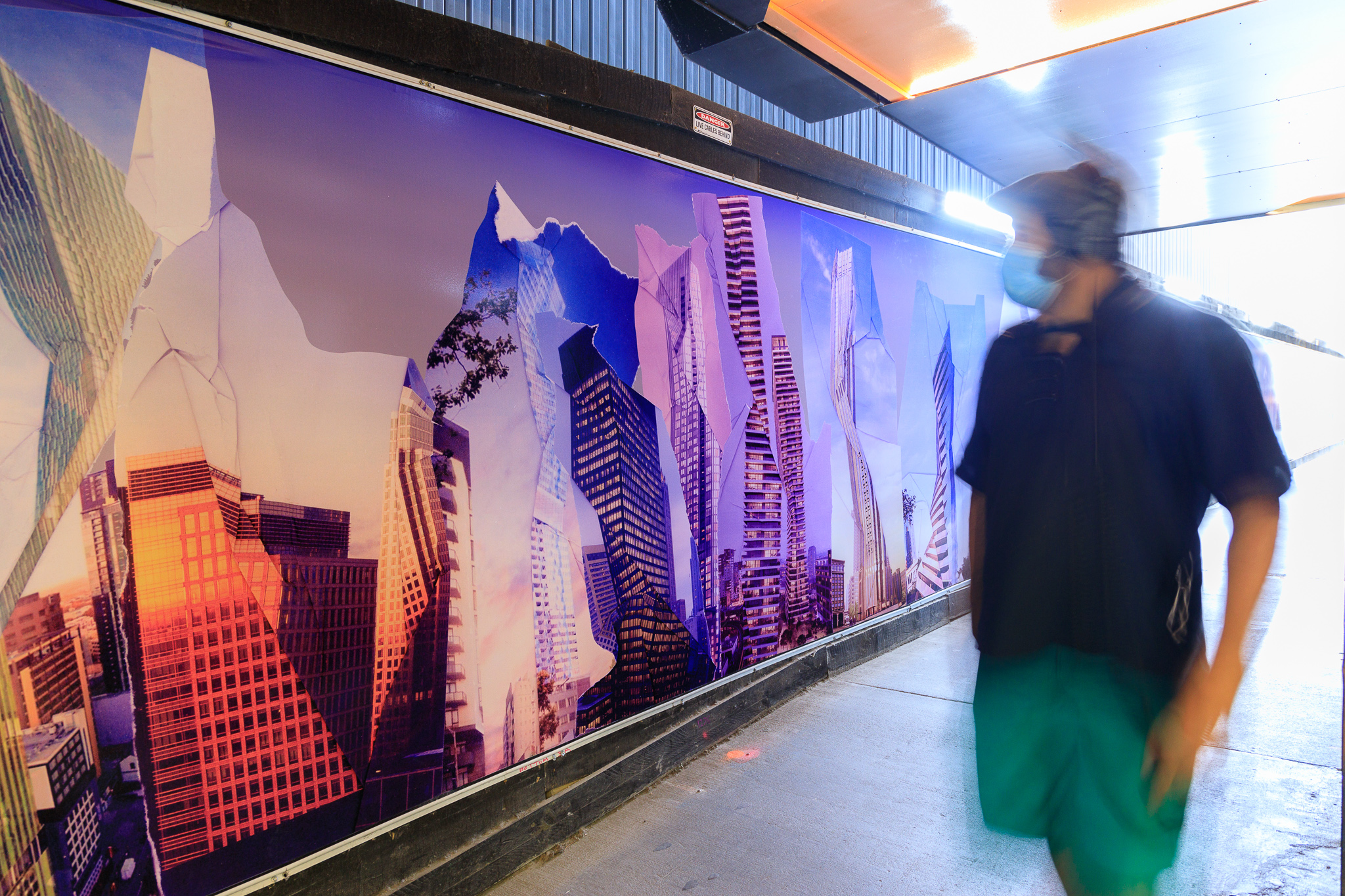 A man in a maks walks past altered photographic images of buildings and mountains, on a wall 