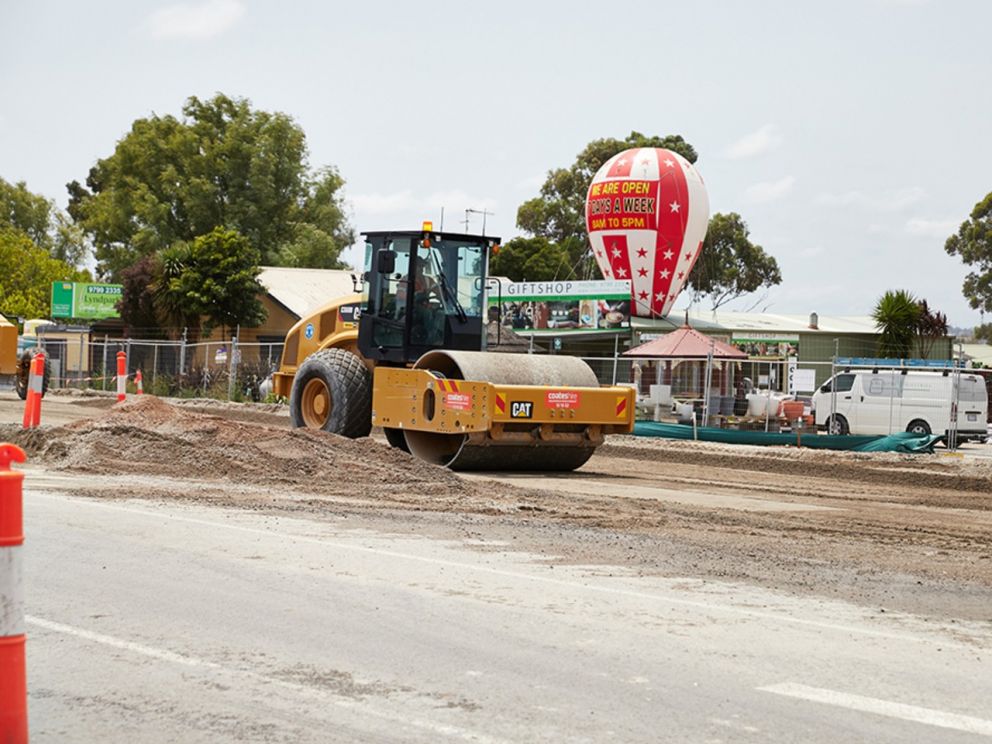 Image of a construction vehicle flattening the road with a strip of shops in the rear