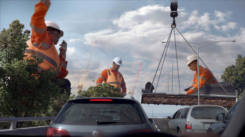 Three giant workers in hi-viz clothes and hard-hats supervise a section of road being lowered into place by a crane. Several cars wait in the foreground.  
