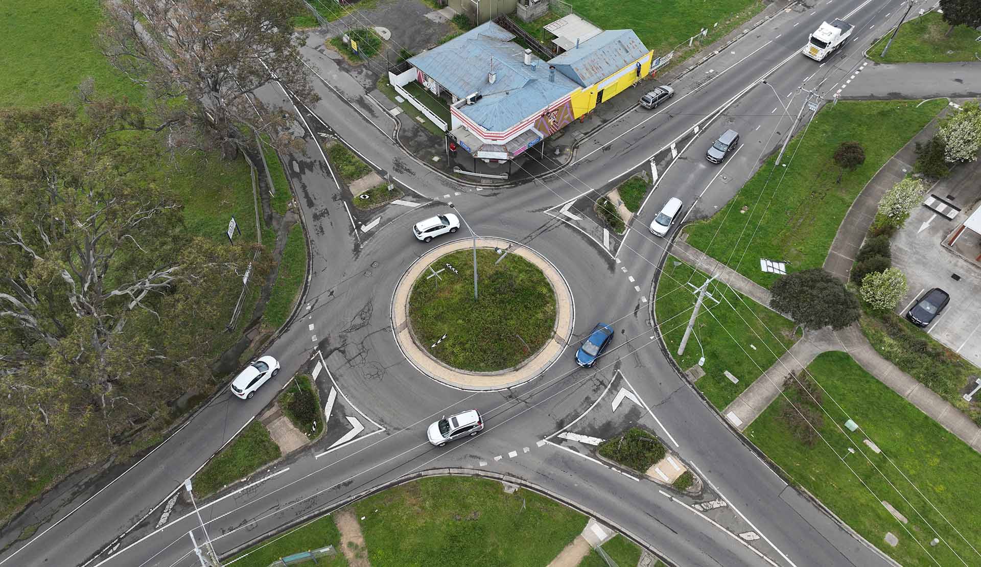 Image of a roundabout within the Yan Yean Road upgrade. The image has shopping strip and bare land surrounding the roundabout. There are cars driving on and off the roundabout