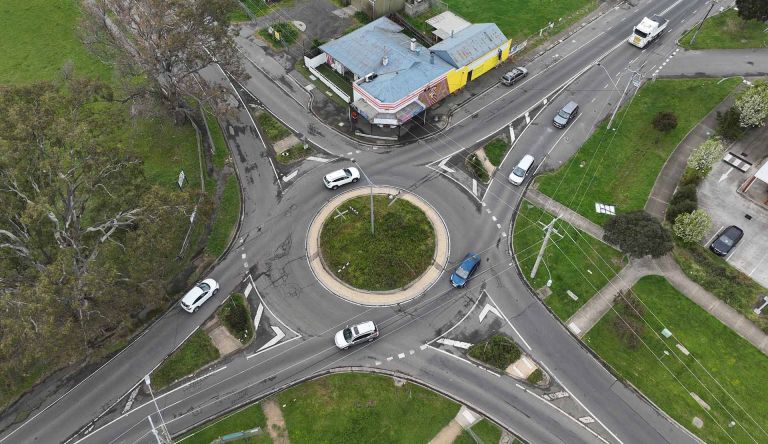 Image of a roundabout within the Yan Yean Road upgrade. The image has shopping strip and bare land surrounding the roundabout. There are cars driving on and off the roundabout