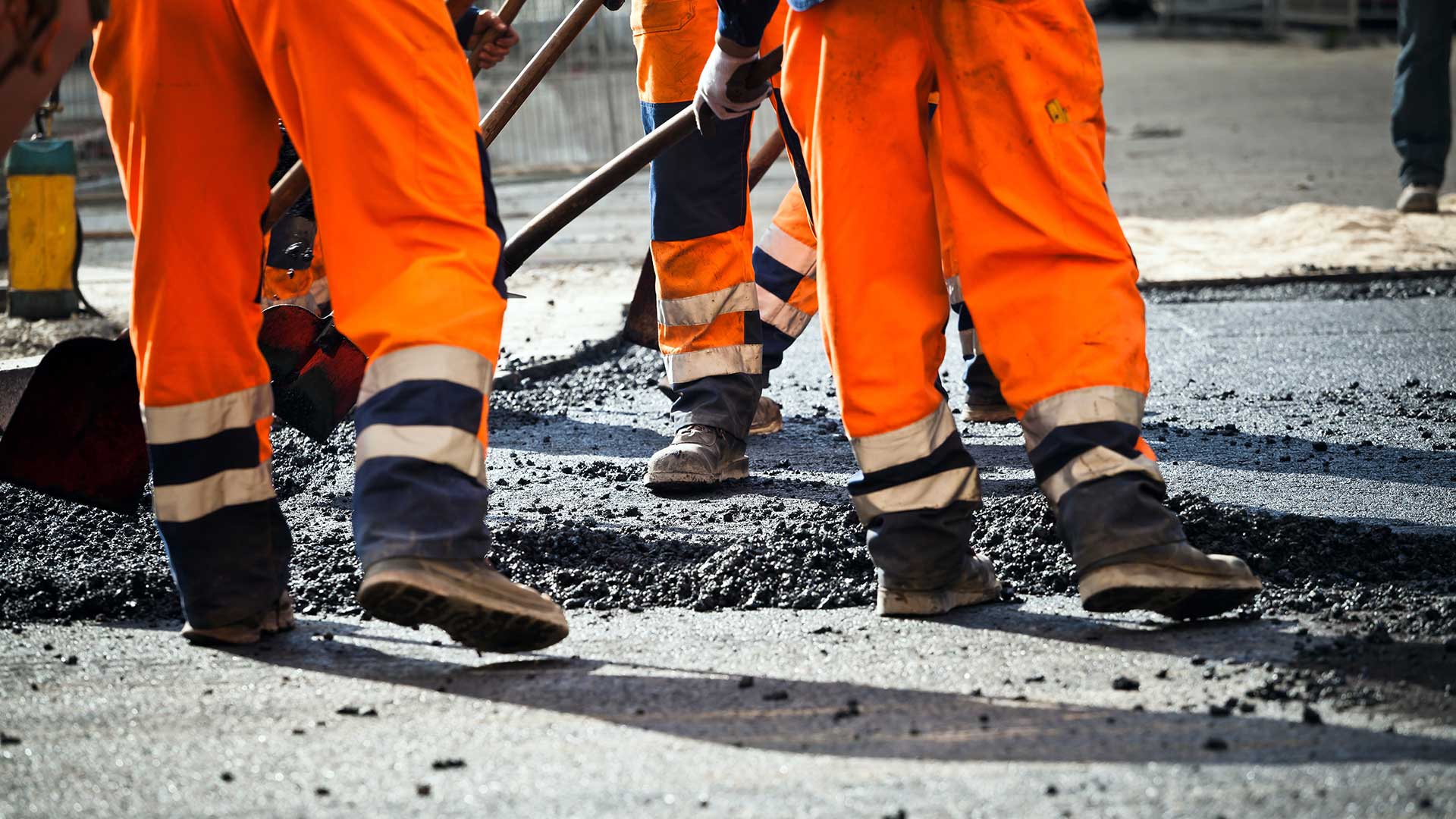 A group of construction workers wearing orange protective gear laying asphalt on a road.