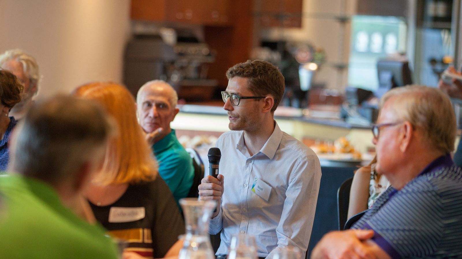 A group of people sitting around a table. The camera is focused on a man wearing glasses and speaking into a handheld microphone.