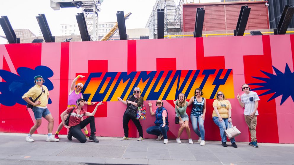A group of people pose in front of a large artwork on a hoarding with the typography ‘Community’.