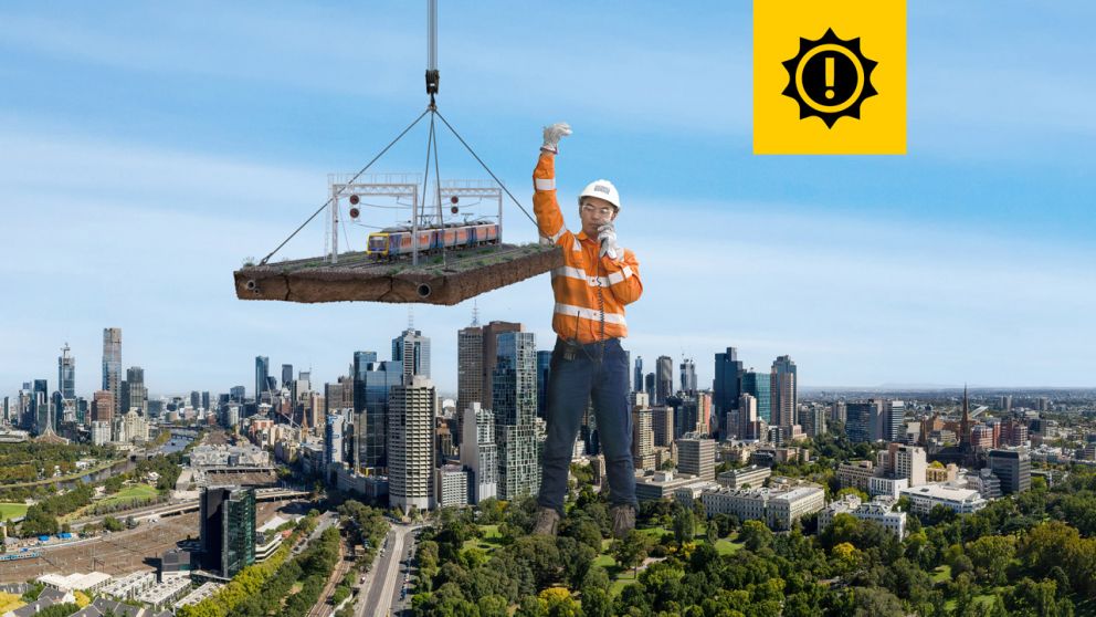 A giant-sized man in a hard hat and work wear stands to the east of the Melbourne CBD, helping to manoeuvre a segment of train track, which is suspended from a crane. 