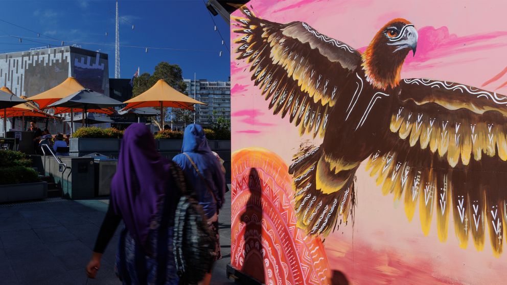 A painted image on a hoarding wall of a large bird next to people walking through Fed Square