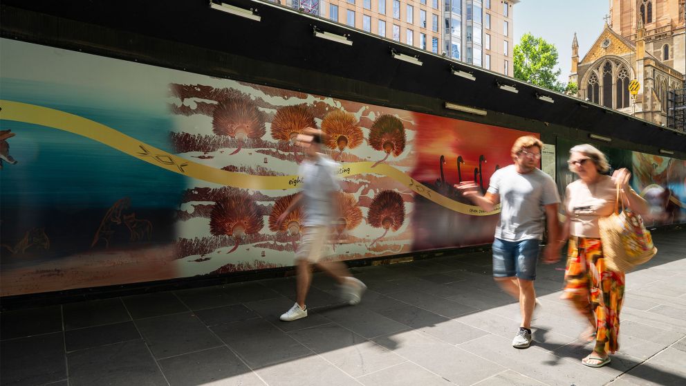 Three people walking in front of colourful construction hoarding that features eight echidnas eating