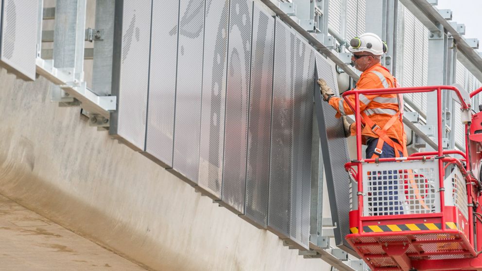 A worker installing a panel of artwork on the Surf Coast HIghway rail bridge. 