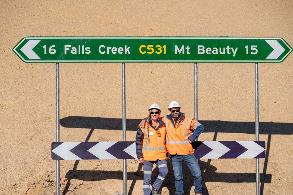 Team members pose for a photo in front of a road sign indicating 16 km to Falls Creek and 15 km to Mt Beauty