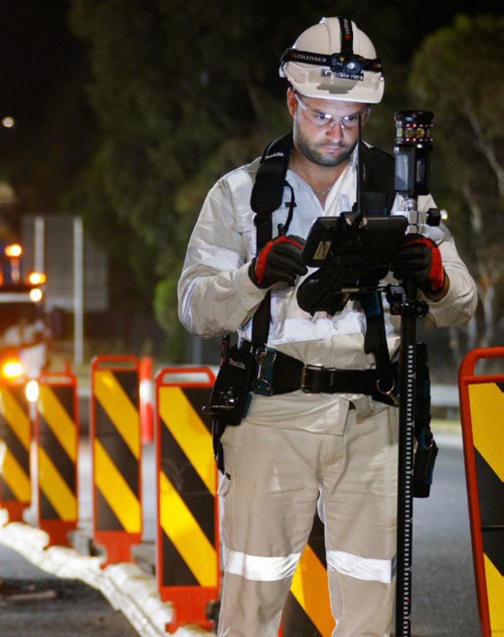 A construction worker in safety gear operates equipment at night near red and yellow barriers on a roadway.