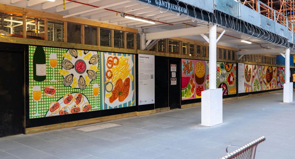 An artwork featuring an assortment of colourful foods on patterned tablecloths installed on a construction hoarding