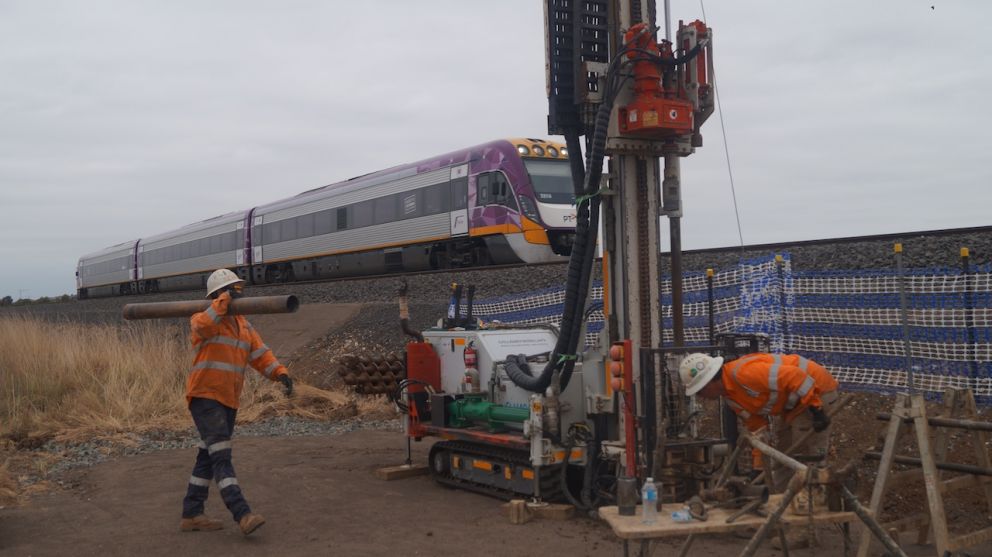 Two workers in orange hi vis, one carrying a pipe and one working near a piling rig.