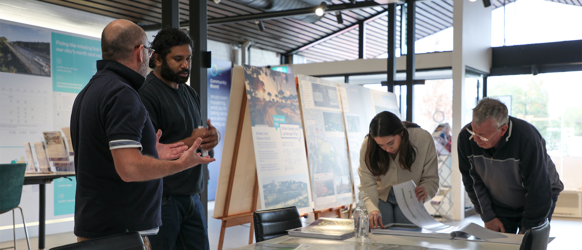 Four people in a modern, well-lit room are gathered around a table covered with maps and documents. Two individuals stand on the left discussing, while two on the right examine papers closely. Behind them, large posters on easels display urban planning or development project information. The setting reflects a collaborative planning session.