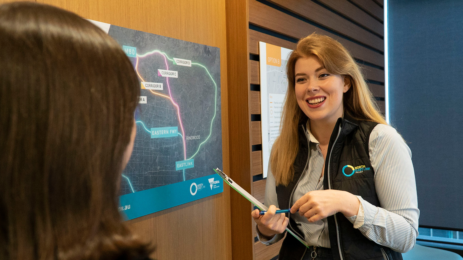 A North East Link Project employee holding a clipboard and pen smiling at another person.