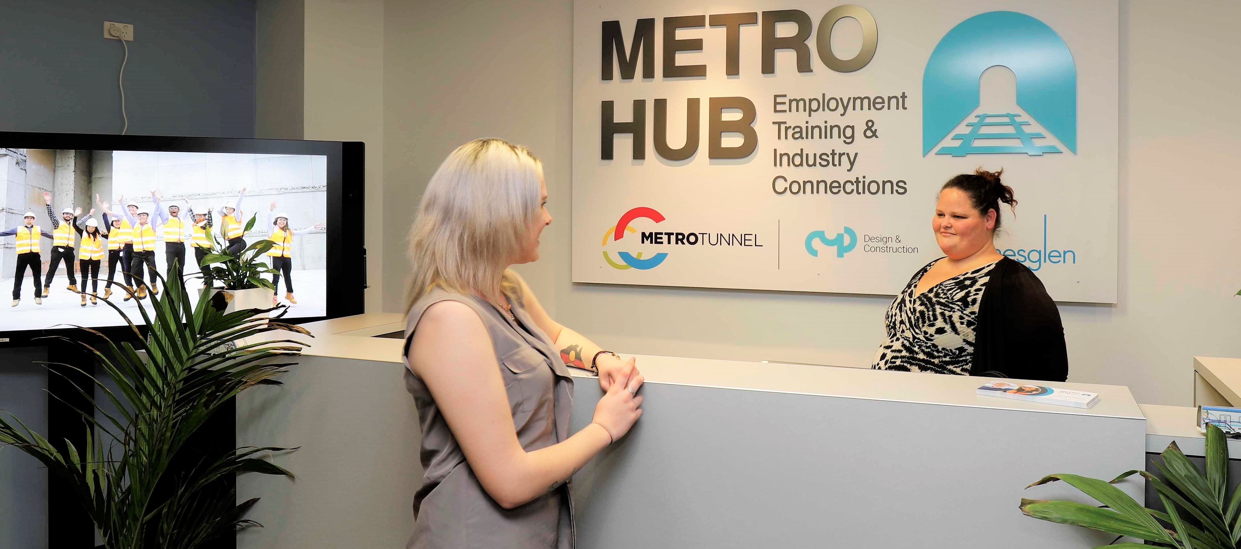 A woman speaks to another woman behind a counter with sign on the wall behind that says 'Metro Hub: Employment Training & Industry Connections