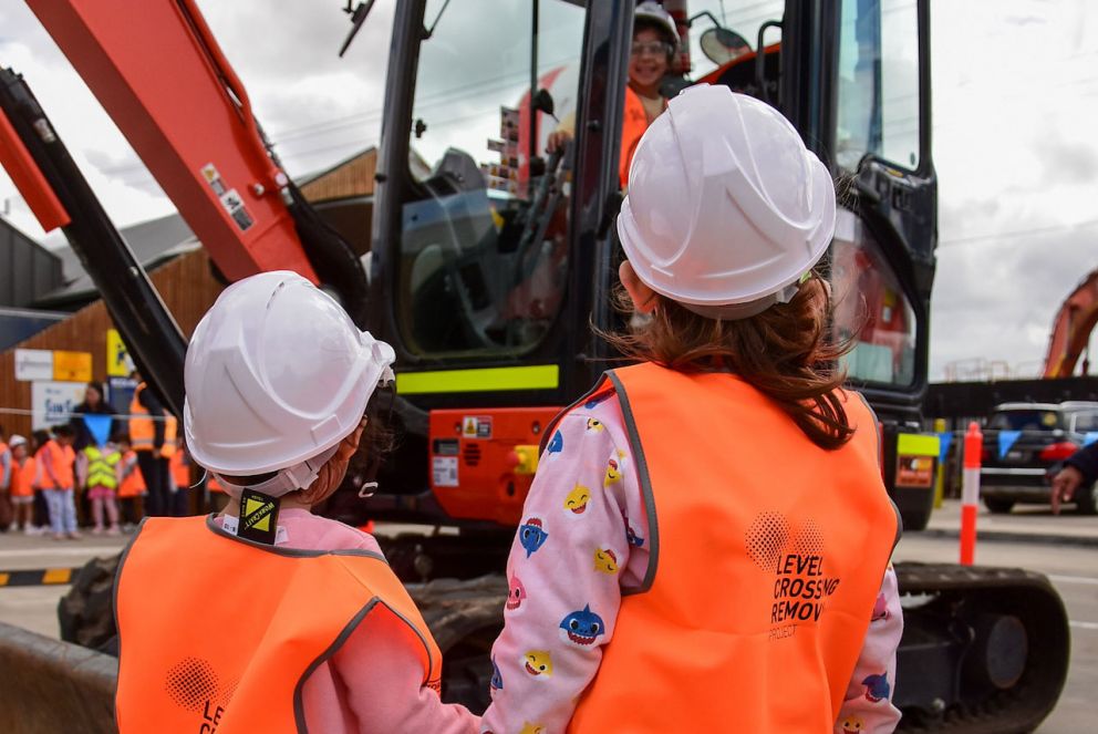 Two children holding hands looking at another sitting in the digger.