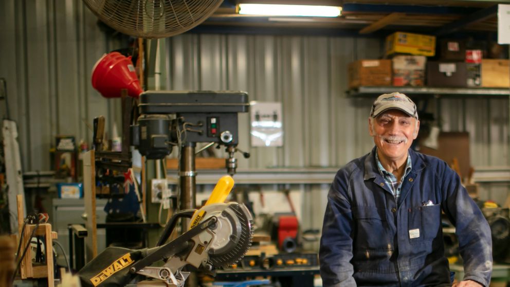 An older man wearing a baseball cap and overalls sitting in a workshop surrounded by tools and smiling.