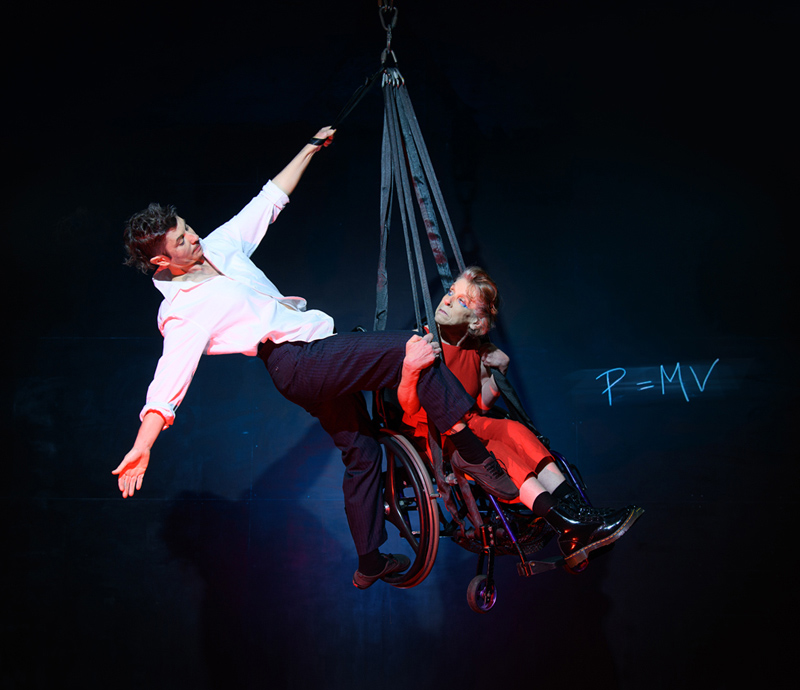 A woman in a wheelchair is suspended in the air with a man being held by her also suspended posing.