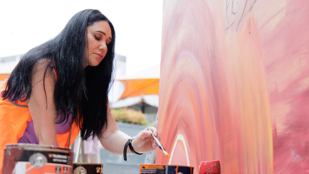 A woman wearing a bright orange vest paint a section of an artwork on a wall