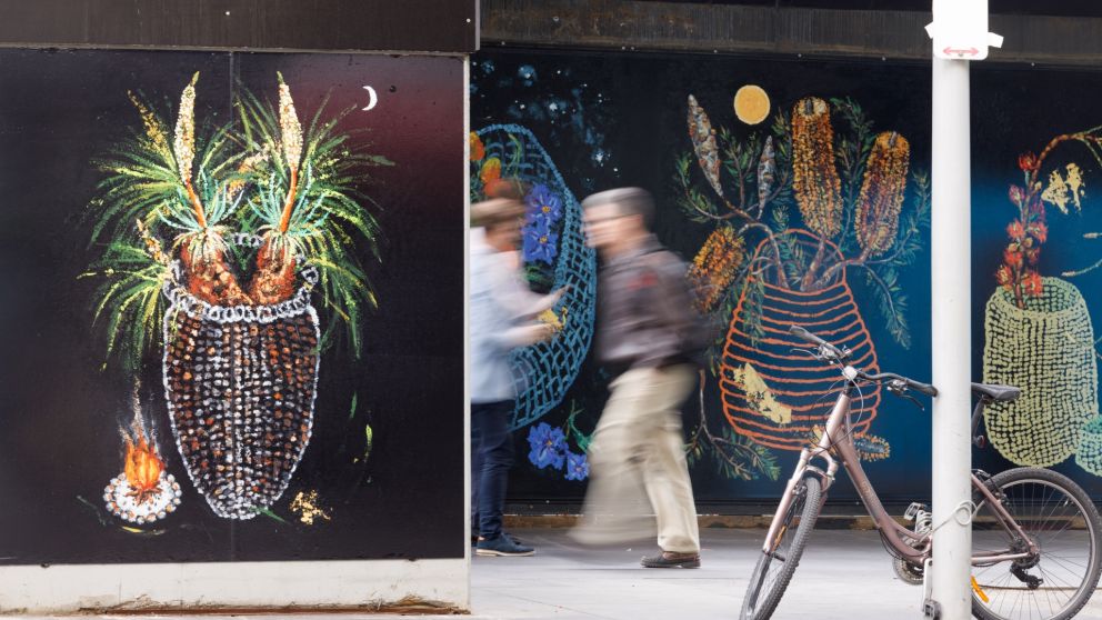 Pedestrians walking past a painting of mutiple hand woven baskets with colourful flowers