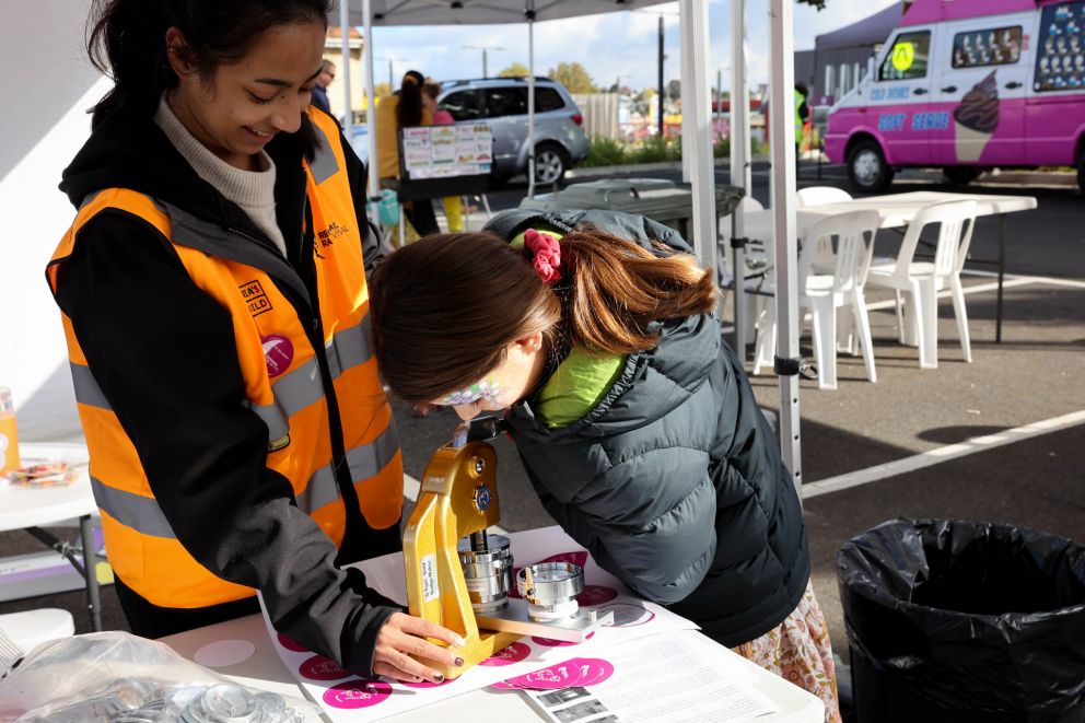 Person using a badge making machine on table with assistance from staff member in high vis vest